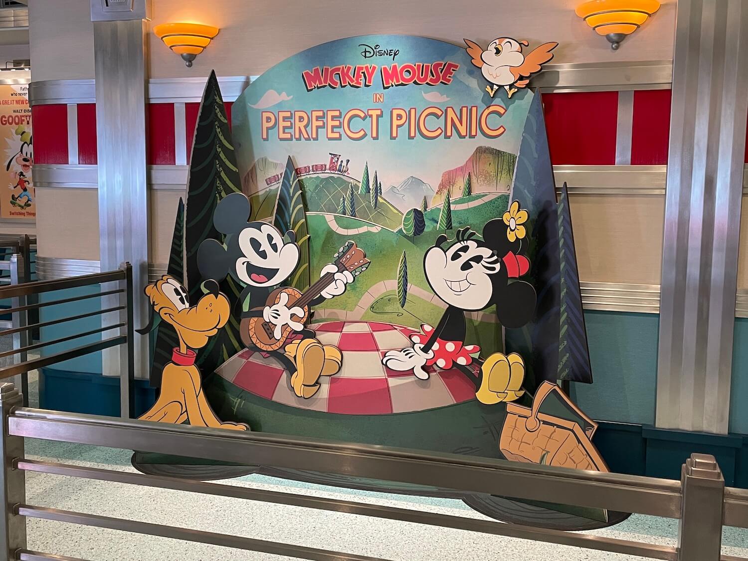 A flat, movie theater-style display at the entrance to the ride, which is also themed like a movie theater. It's Mickey, Minney, and Pluto on a picnic blanket in a verdant park. Above them it says “Mickey Mouse in Perfect Picnic,” and we see Goofy conducting a steam train on the horizon.