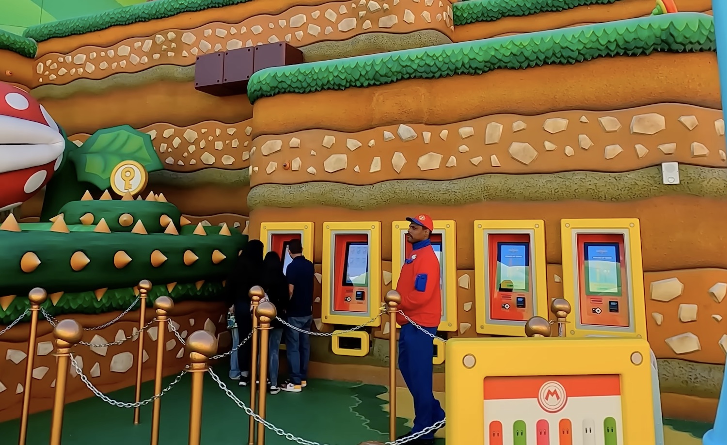 A few people stand around one of the five Power Band kiosks that are built into a wall. There's a bored employee guarding the mushroom stanchions.