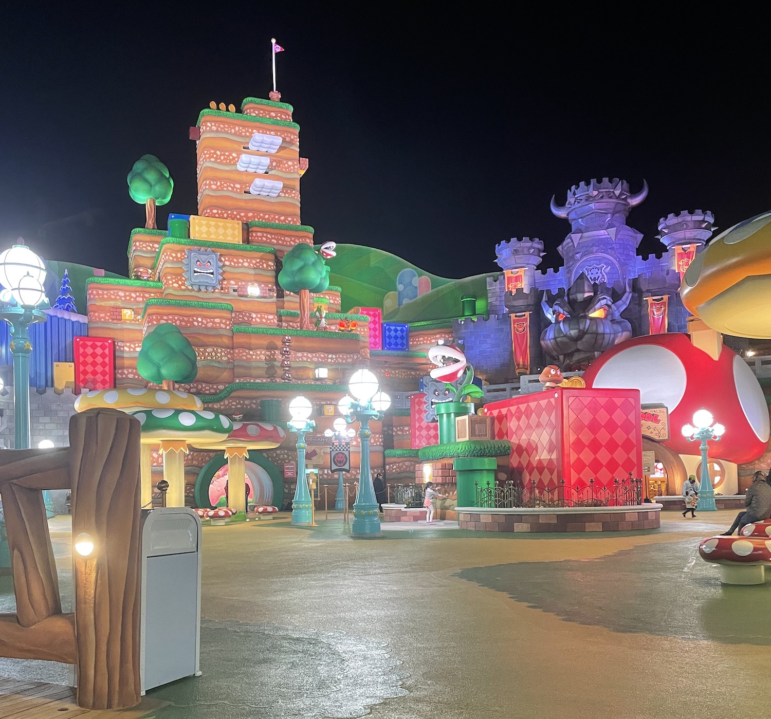 One of the big set pieces at Super Nintendo World at night. We see Bowser's castle, a big piranha plant, and a tall mountain covered with moving platforms and a flag at the top.