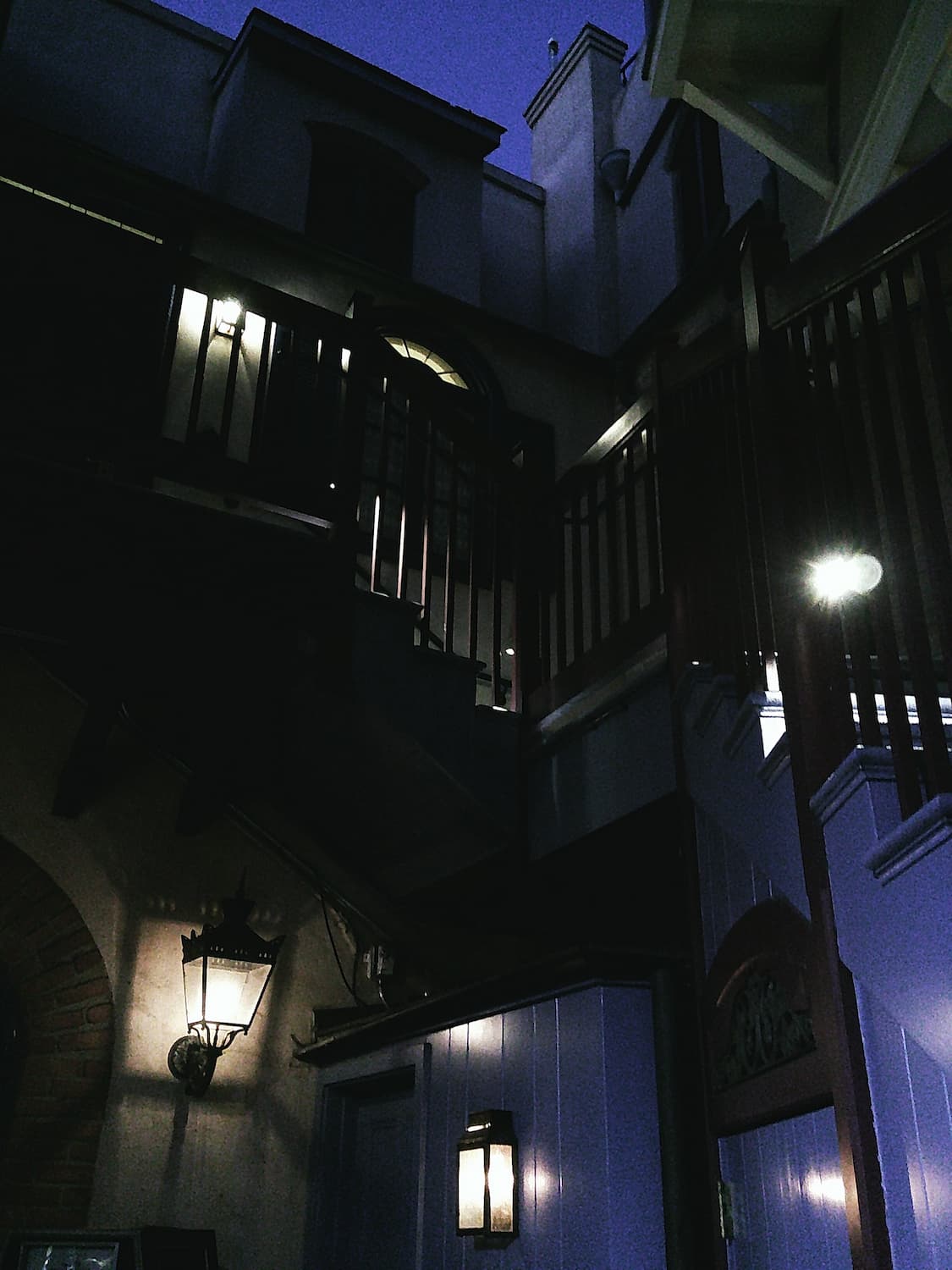 A similar photo of a different location, this time at dusk. A stairway leads up the interior edge of a small courtyard, shiplap panels and wooden railings in line with the sharp edges of a multifaceted structure. Iron-grated lanterns illuminate the scene from many directions.