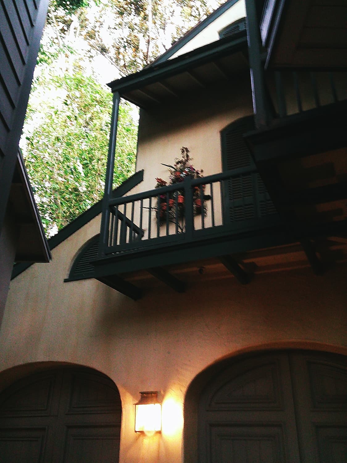 Looking up at the exterior of a stucco building to a second-story balcony. Dark railings match the dark trim of the roofline, and a simple floral arrangement rests in a niche.