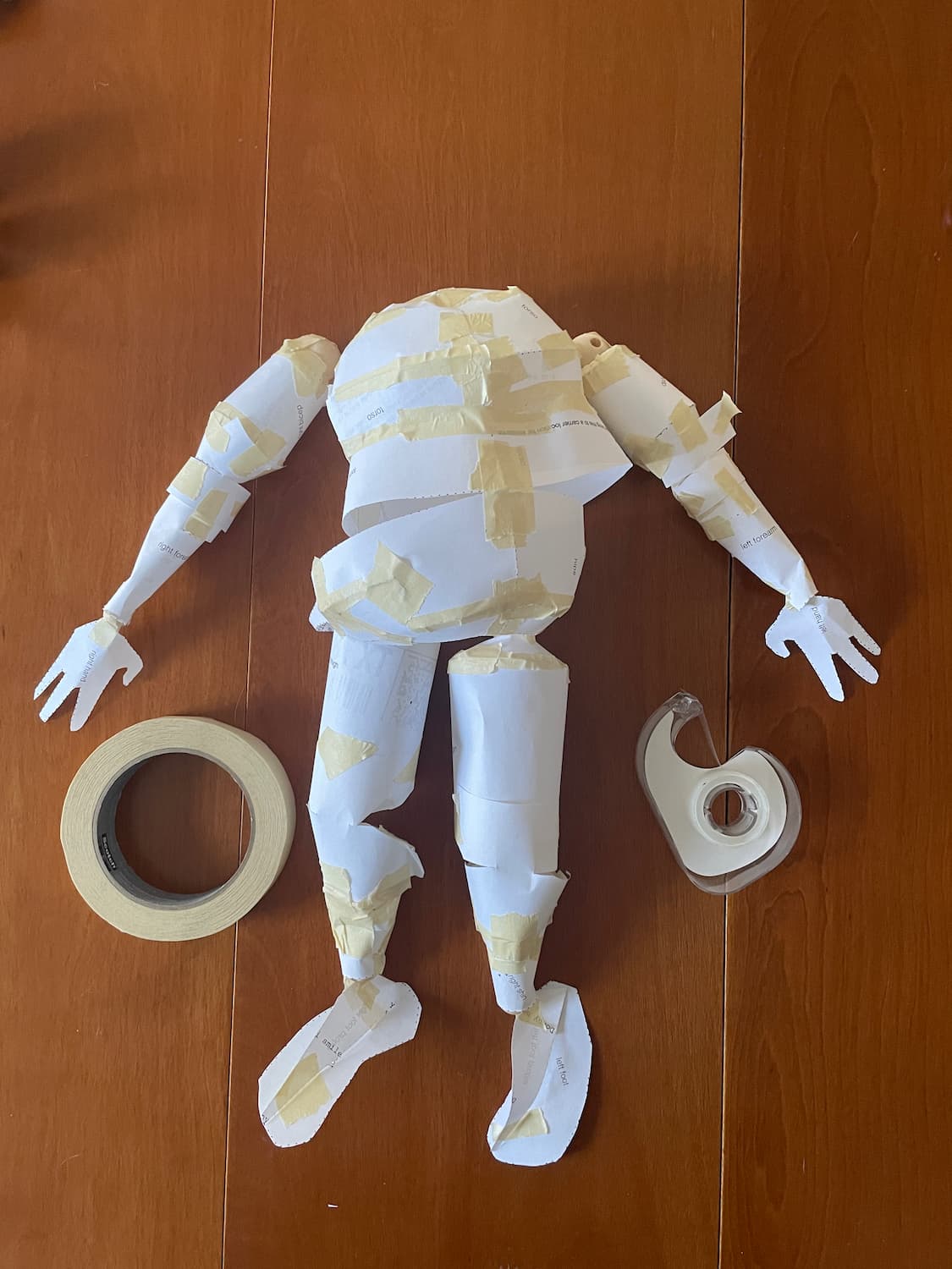 A human figure lying flat on a wood table, cut from copy paper and taped into geometrically-simplified blobby shapes. It bears a striking resemblance to the 3D render from earlier.