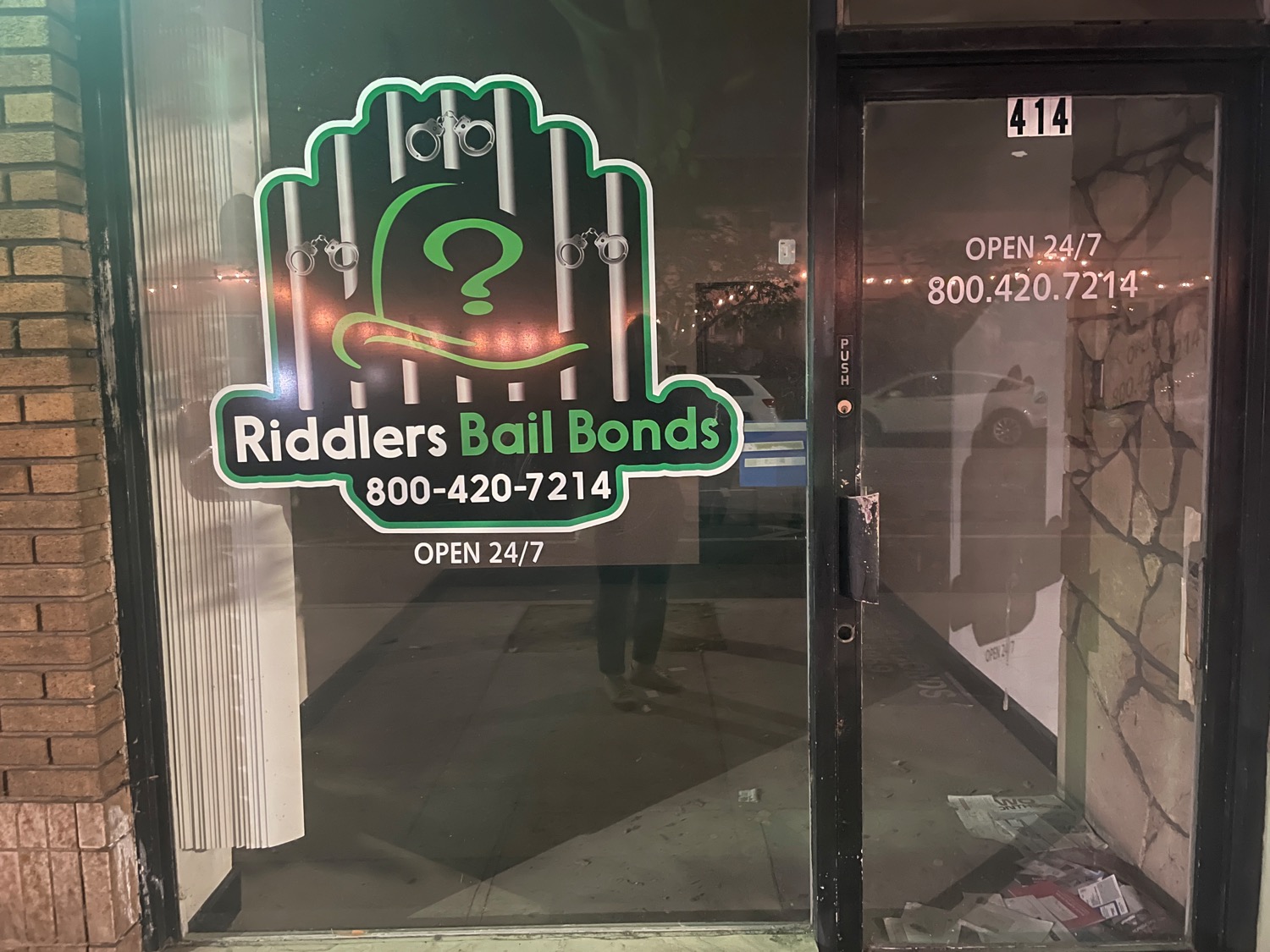A small storefront that is completely empty. The door and window include text and graphics that read RIDDLERS BAIL BONDS, OPEN 24/7