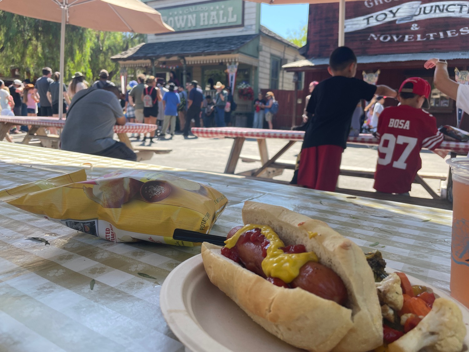 A first-person view of a hot dog on a paper plate. In the background, modern-timey guests gather around an old-timey storefront.