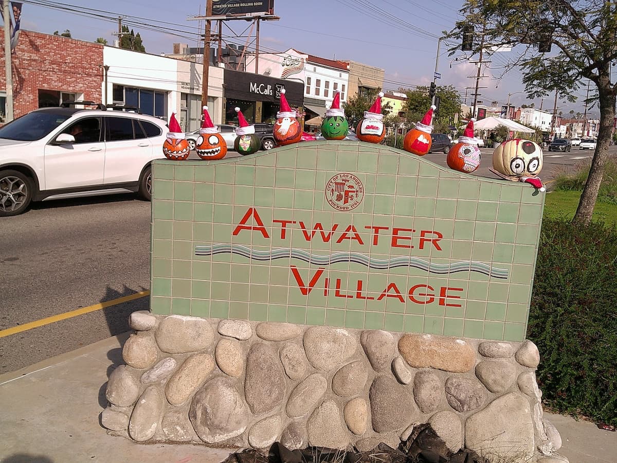 A dozen or so pumpkins with painted faces sitting atop a sign that says Atwater Village, positioned on the median of Glendale Boulevard