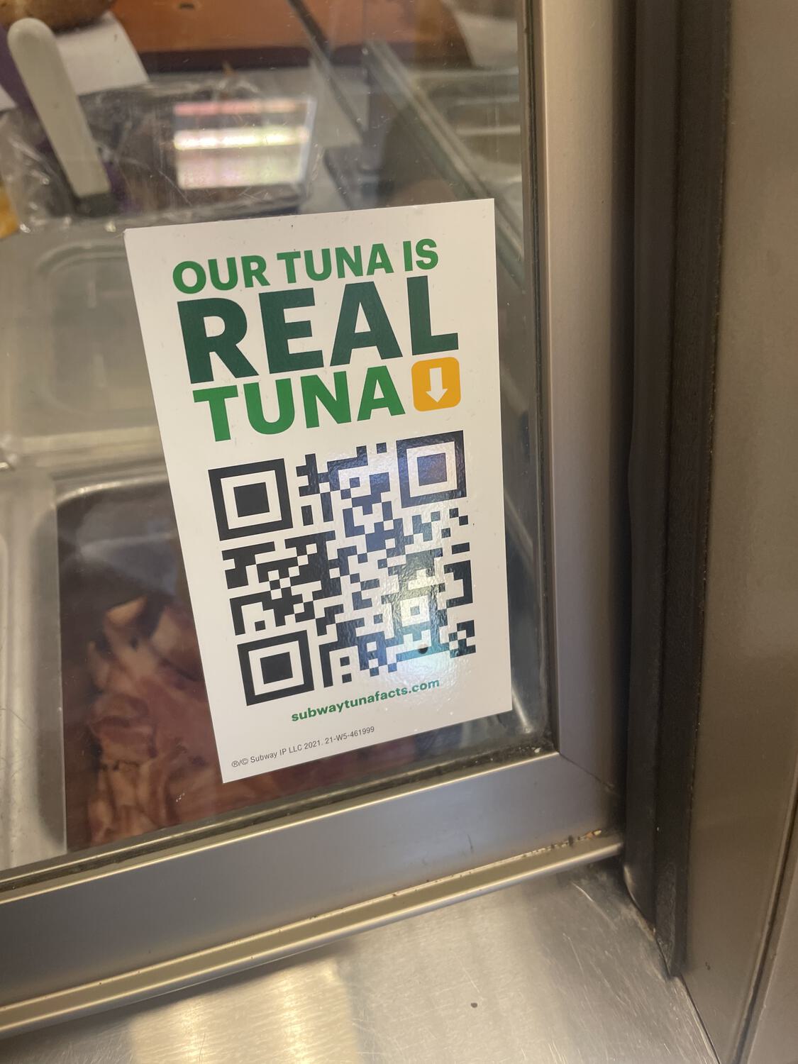 A sticker on the front glass of a Subway display case that reads "Our tuna is real tuna" with a QR code