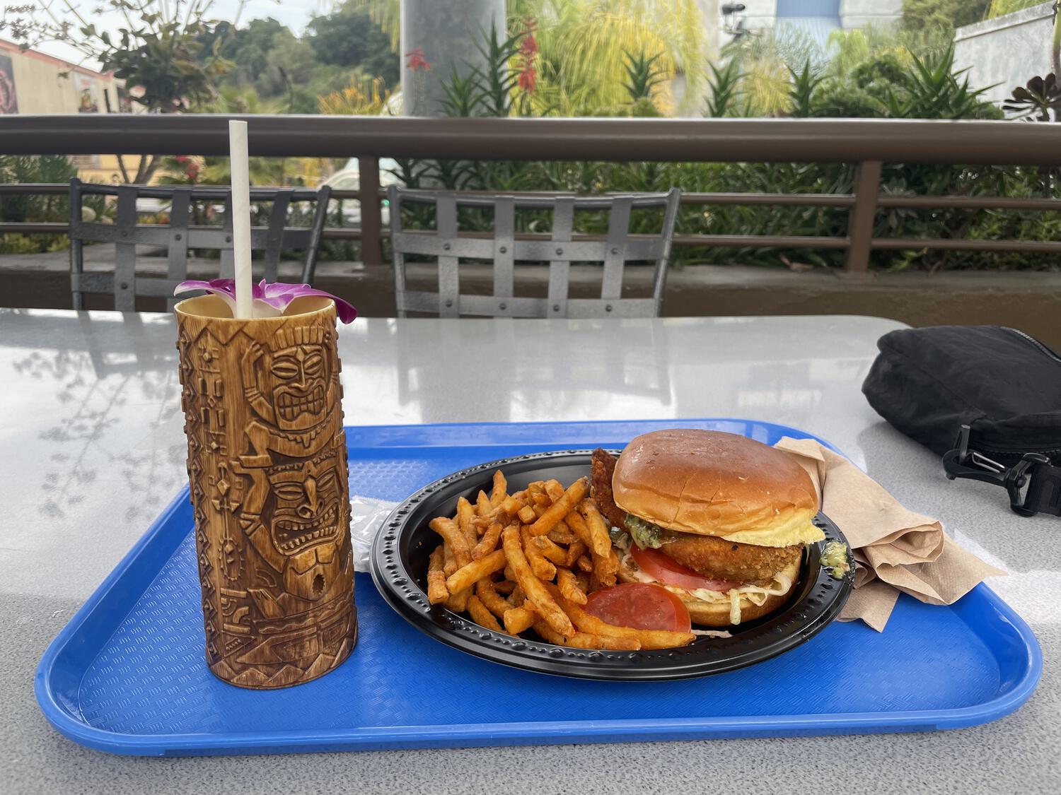 A chicken sandwich and fries with a tall tiki cocktail, all on an outdoor table with foliage and soundstages in the background