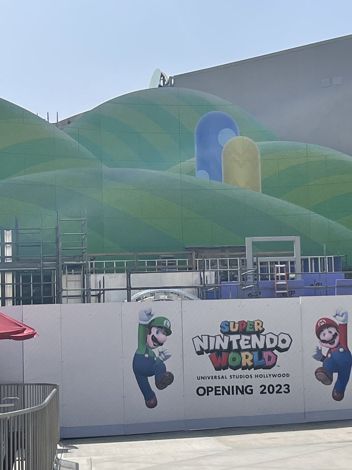 A construction wall reading "Super Nintendo World / Universal Studios Hollywood / Opening 2023," with unfinished green cartoon hills and scaffolding rising behind them