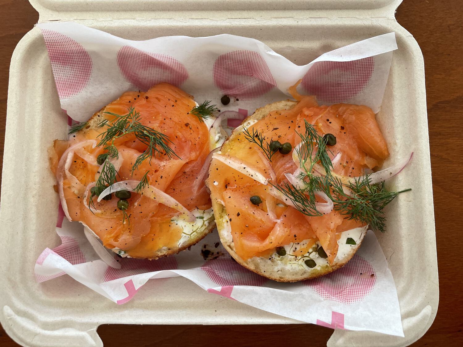 A bagel with lox, capers, dill, onions, and cream cheese, artfully presented in a to-go container