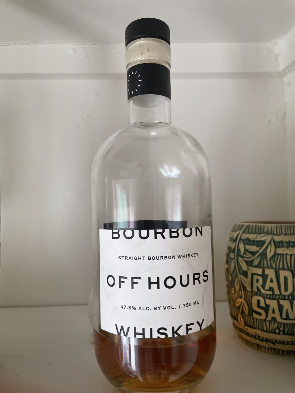 A mostly-empty bottle of Off Hours brand bourbon sitting on a shelf beside a Trader Sam's tiki mug