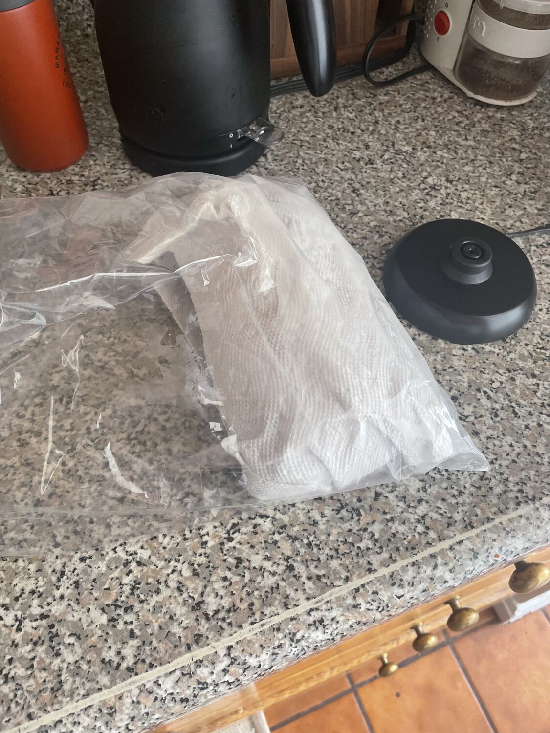 A loaf of banana bread wrapped in paper towel and plastic wrap, sitting on a cluttered kitchen counter