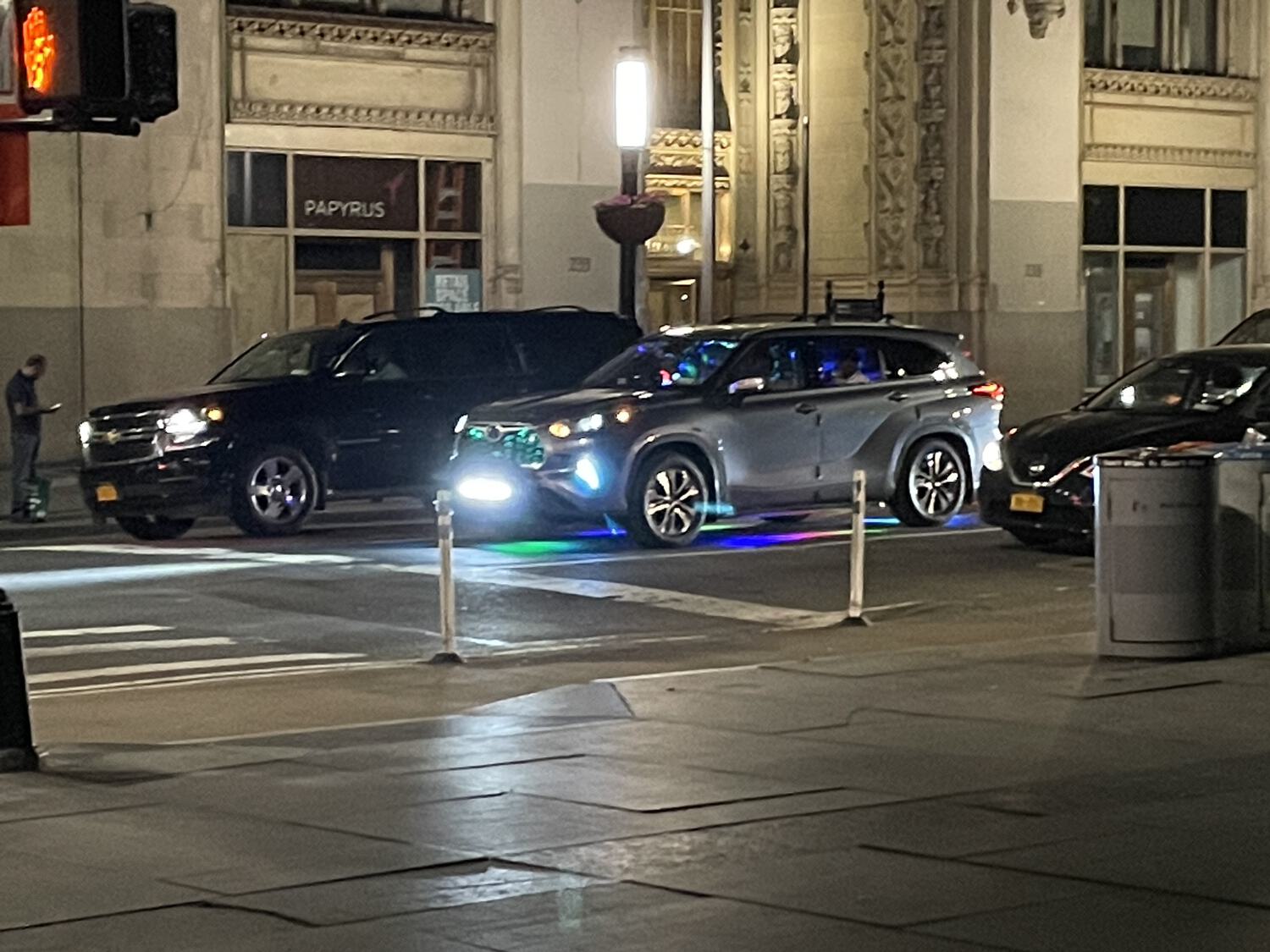 A zoomed-in shot of a car stopped at a red light. There are colorful party lights inside the car, as well as illuminating its grill and the road beneath it.