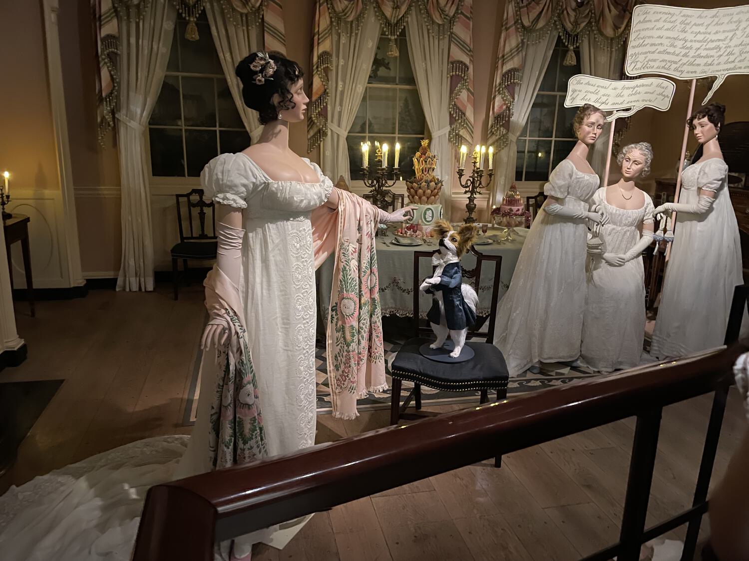 Mannequins in fine 18th Century eveningwear gather in a period room. There are illegible speech bubbles above a group that are watching another from afar. The solo mannequin is playing with a dog, standing upright on a chair, also dressed formally.