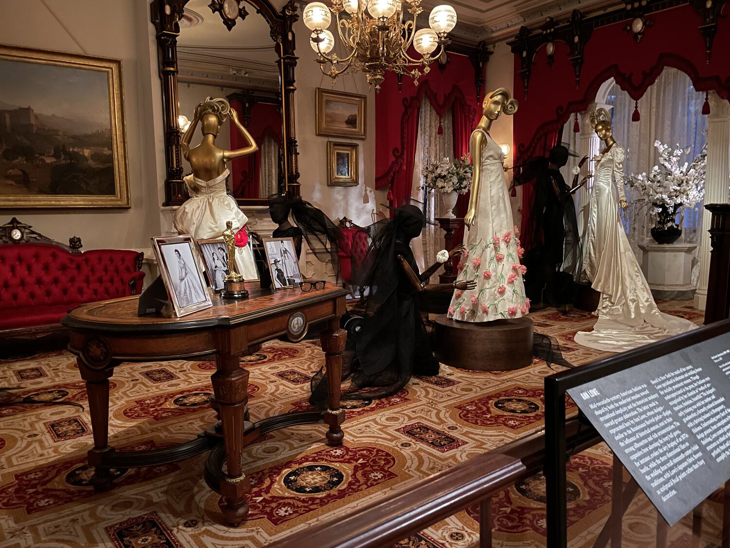Ghostly black mannequins in transparents hats and gowns fit fine eveningwear on gold mannequins amidst Hollywood paraphernalia in an old-fashioned room
