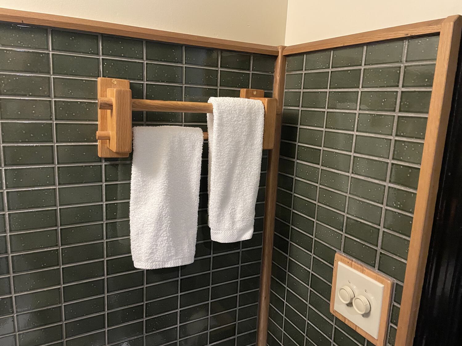 A wooden towel rack with two white hand towels on it, mounted to a wall half-clad in green subway tile. The wall is trimmed with wood matching the towel rack