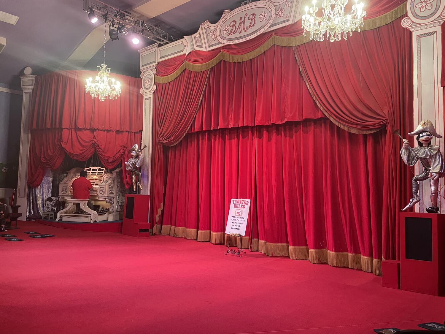 A closed red curtain in a small but finely-decorated theater. An organist plays in the background.