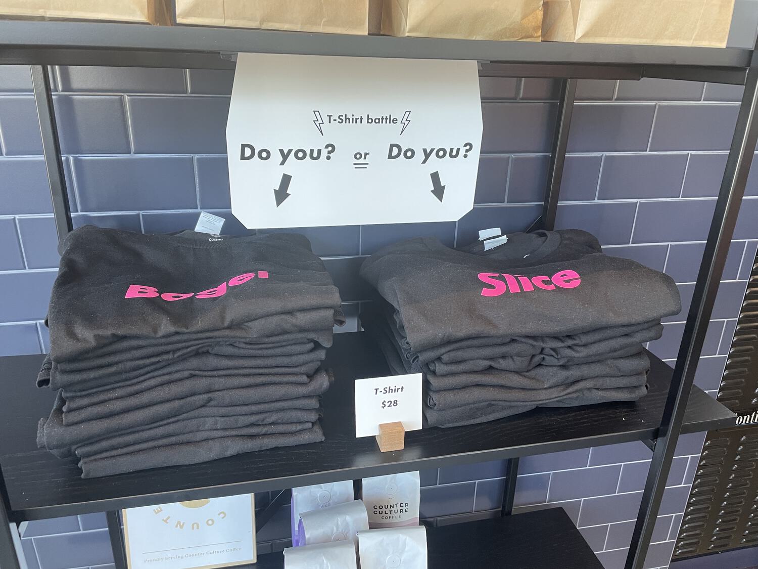 Two piles of t-shirts for sale, one printed with "Bagel" and the other printed with "Slice." Above them is a sign that reads "T-Shirt battle: do you? or do you?"