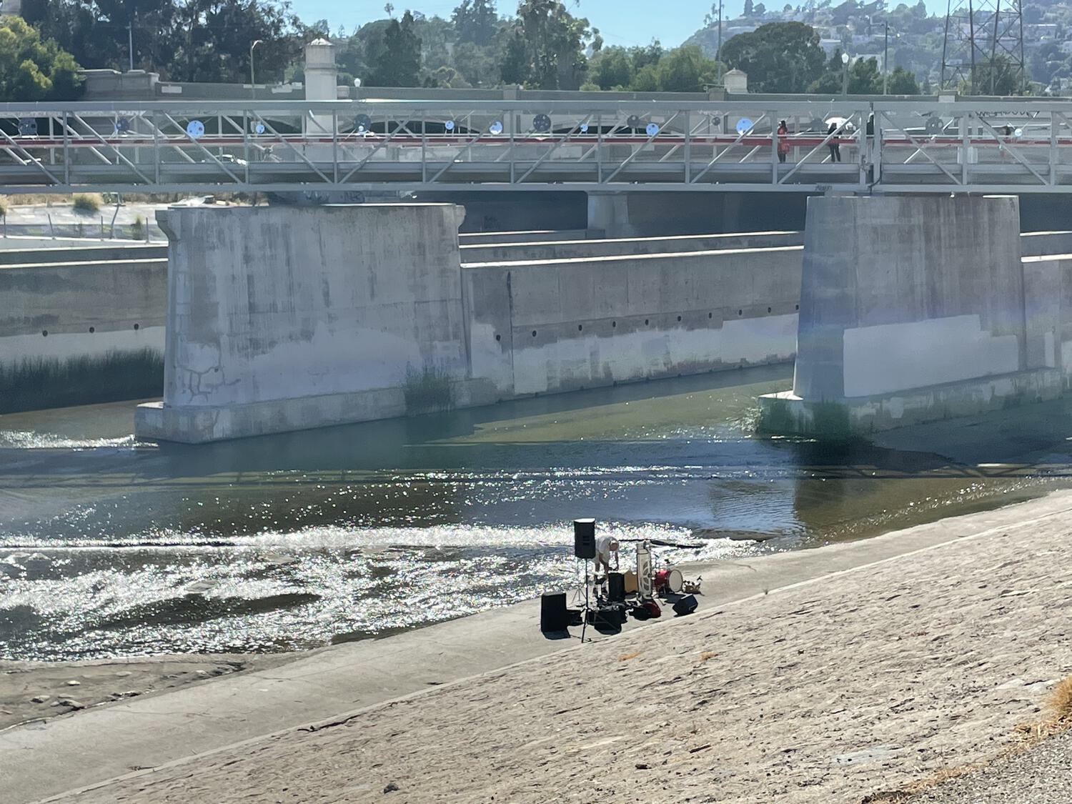 A distant view of an art installation on the Red Car Bridge, as well as a musician setting up equipment on the bank of the LA River