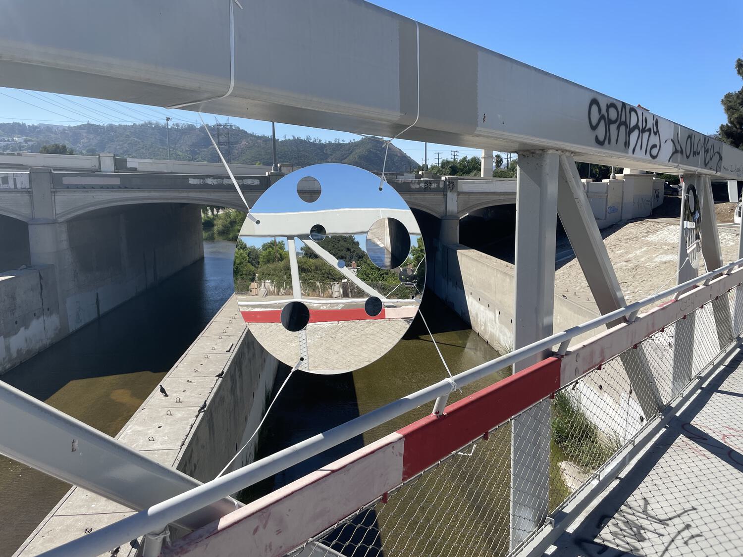 A reflective circle with smaller circles cut out of it, suspended in the air along the railing of a bridge over the LA River
