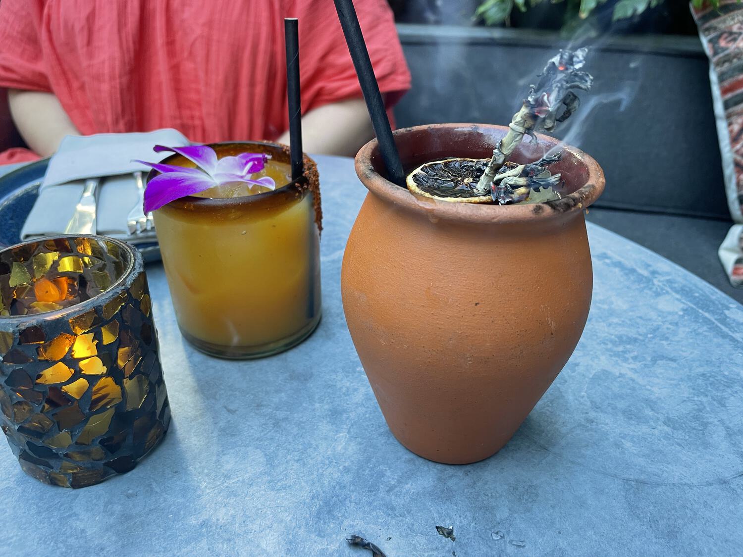 Two cocktails on a restaurant table: one with a hibiscus flower, and one with a burning herb