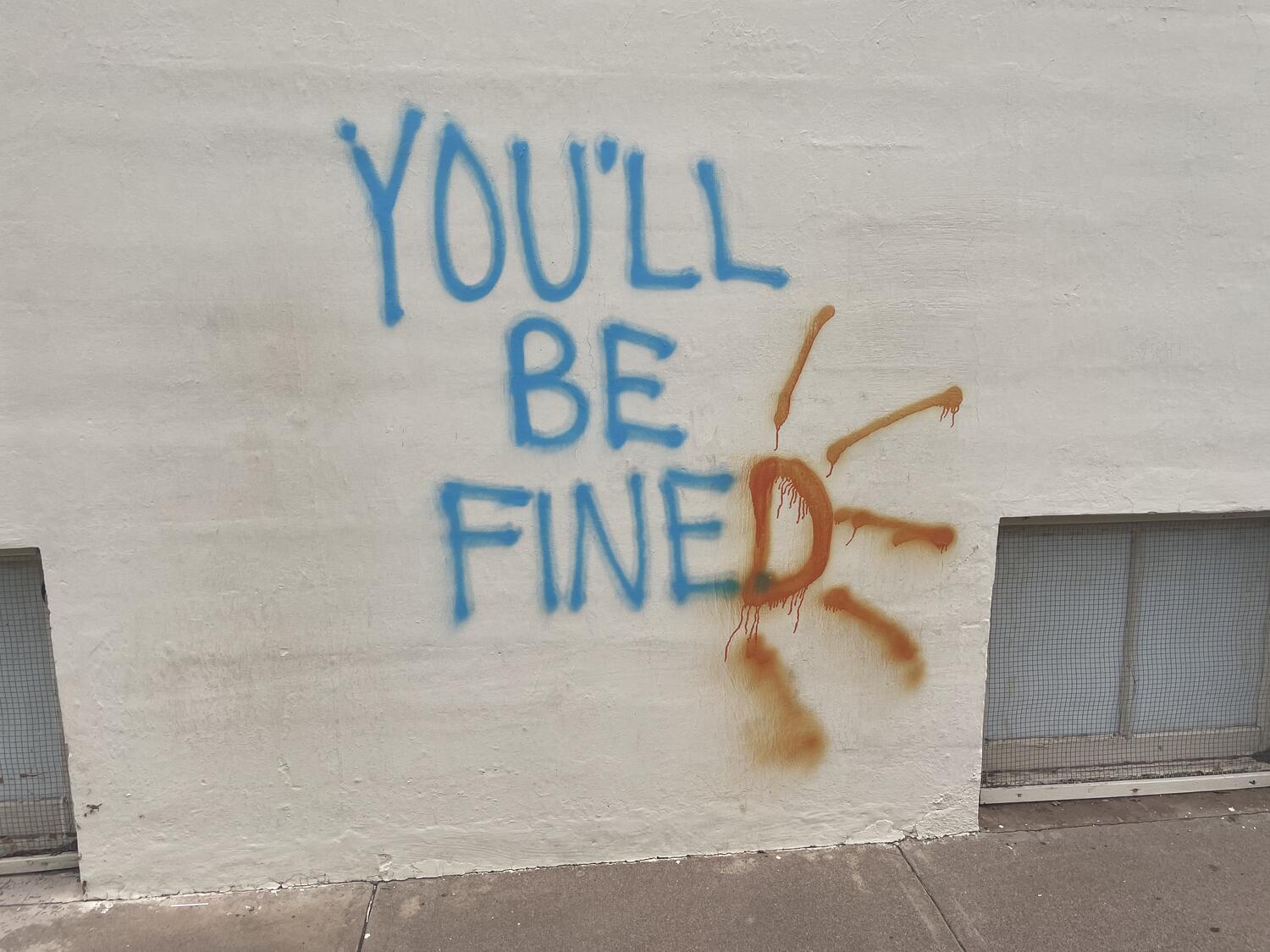 Graffiti that reads “You'll Be Fine” in blue with a “D” in orange at the end so it reads “You'll Be Fined”