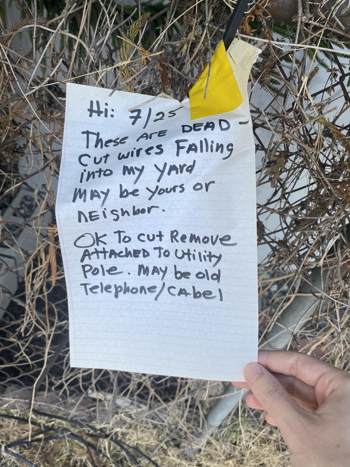 A hand-written note taped to a wire draped over a chain link fence. It reads: “Hi: 7/25 These are dead - cut wires falling into my yard may be yours or neighbor. Ok to cut remove attached to utility pole. May be old telephone/cabel [sic]”