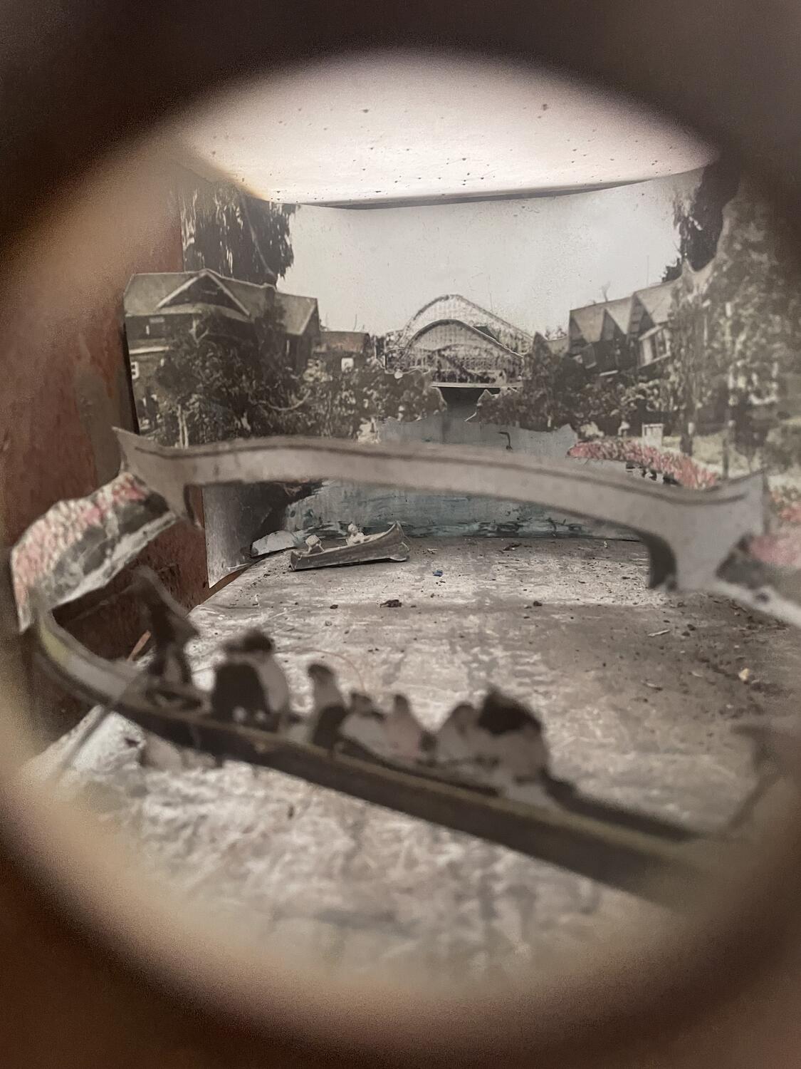 The interior of a shadowbox, seen through a peephole that obscures the borders of the photo. It's like a layered old sepia photograph, bridges and canoes and old houses and a rollercoaster