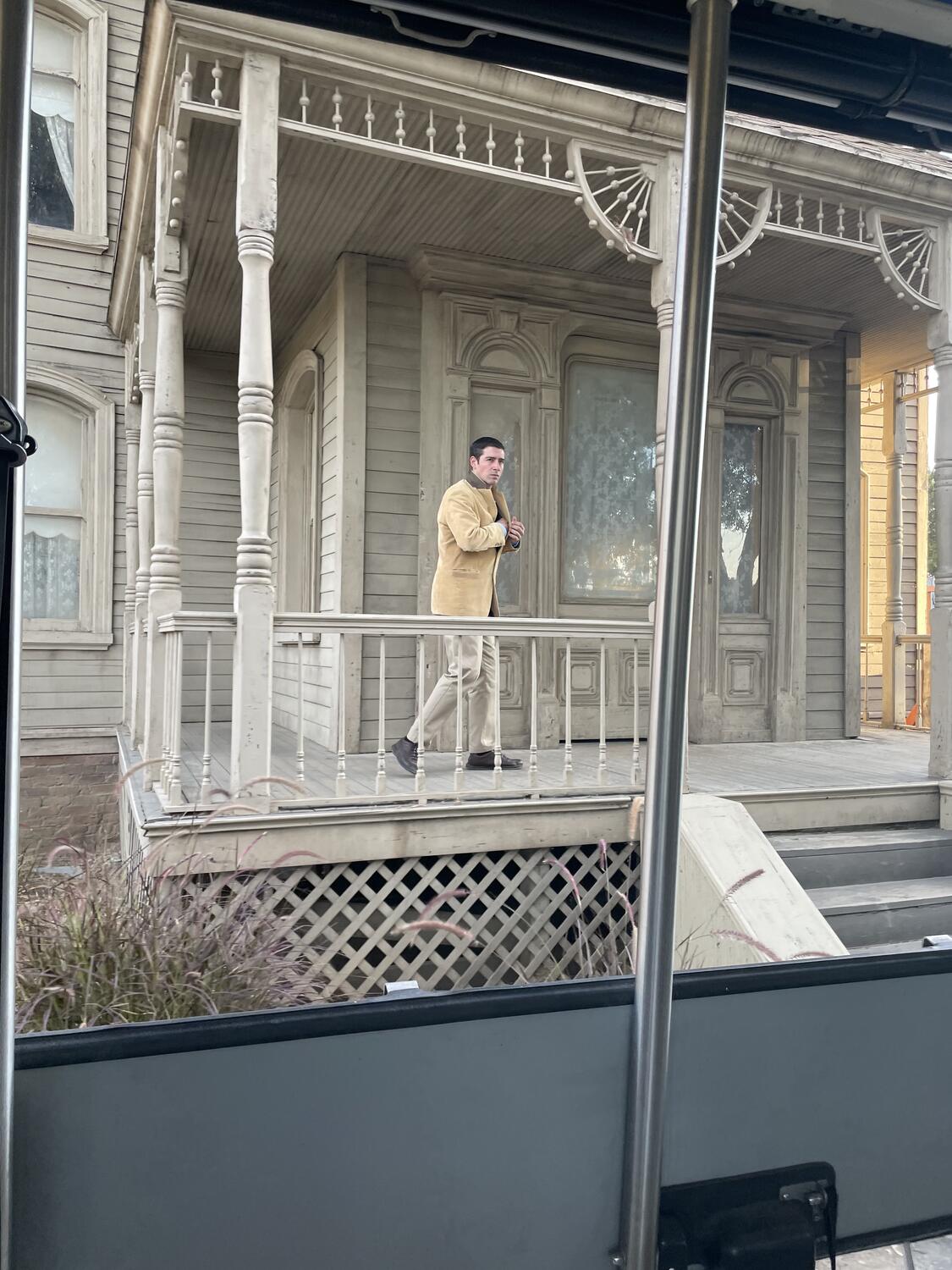 An actor in a tan jacket stands on the porch of an old mansion. He's reaching into his jacket for a big knife.