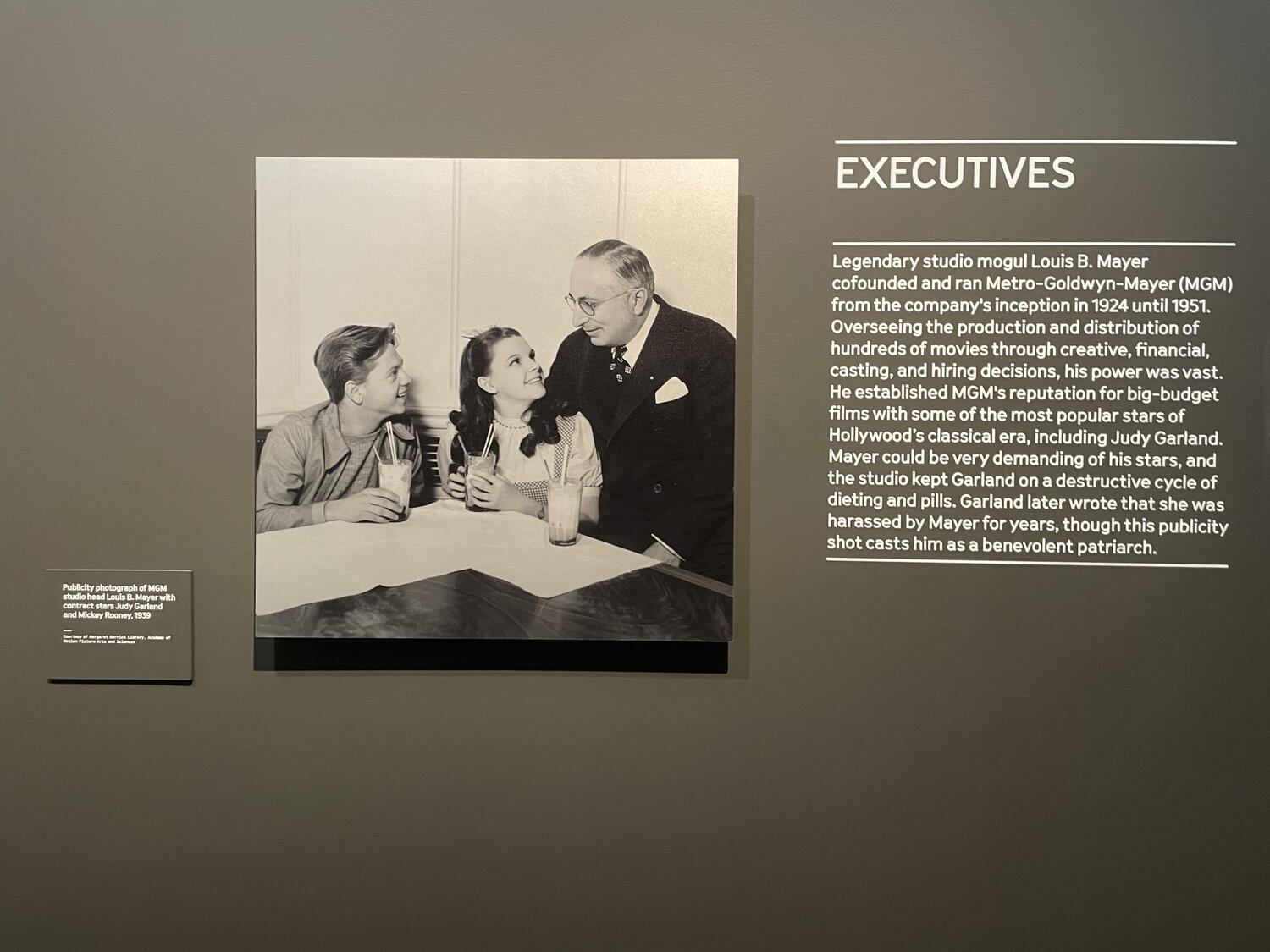 A photo mounted on the wall beside text that reads, “Executives - Legendary studio mogul Louis b. mayer cofounded and ran Metro-Goldwyn-Mayer (MGM) from the company’s inception in 1924 until 1951. Overseeing the production and sitribution of hundreds of movies through creative, financial, casting, and hiring decisions, his power was vast. He established MGM's reputation for big-budget films with some of the most popular stars of Hollywood’s classical era, including Judy Garland. Mayer could be very demanding of his stars, and the studio kept Garland on a destructive cycle of dieting and pills. Galrand later wrote that she was harassed by Mayer for years, though this publiciyt short casts him as a benevolent patriarch.” 