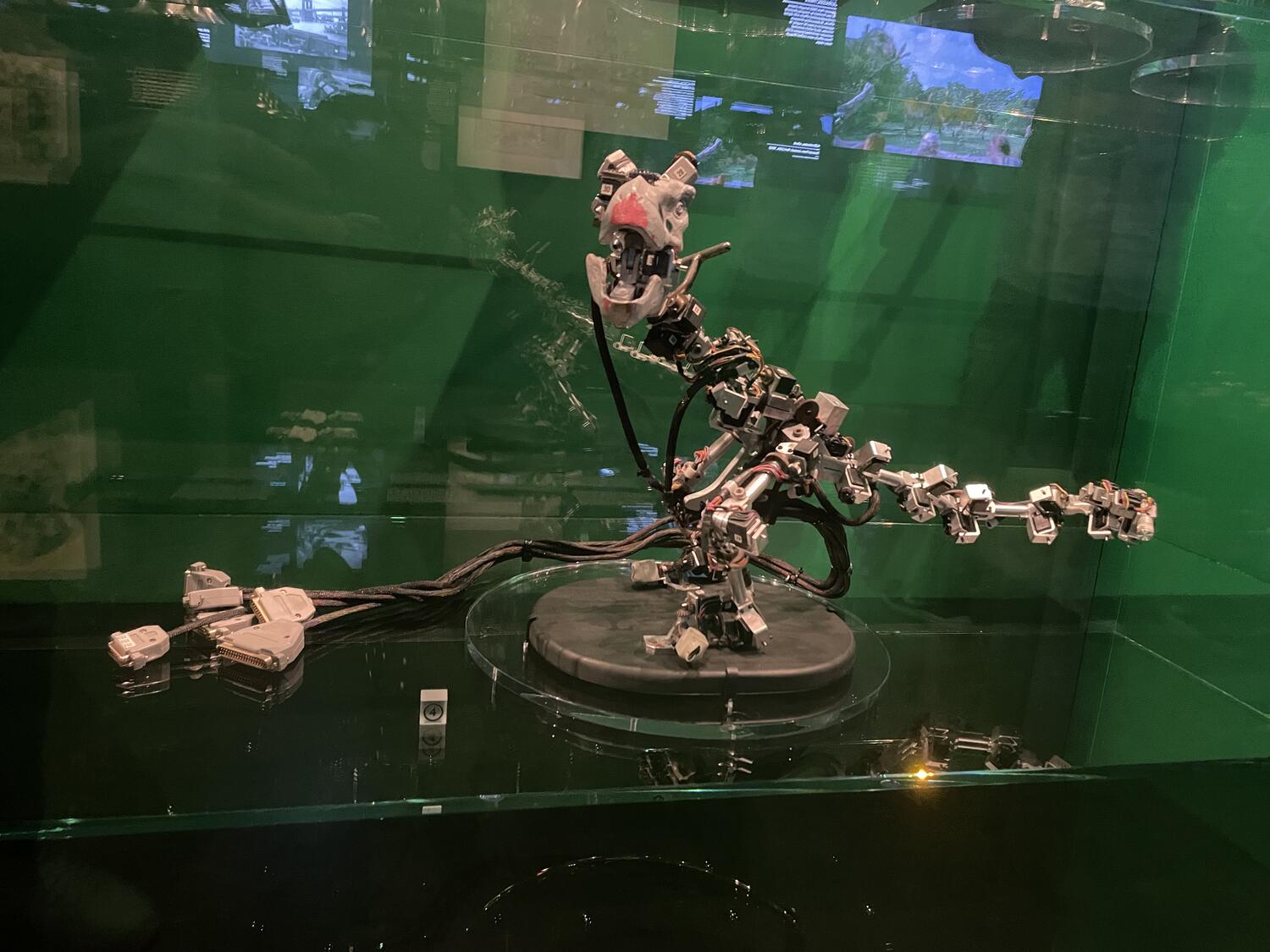 A mechanical dinosaur made of electronic components with some heavy plugs hanging off one side, safely behind glass in a display case