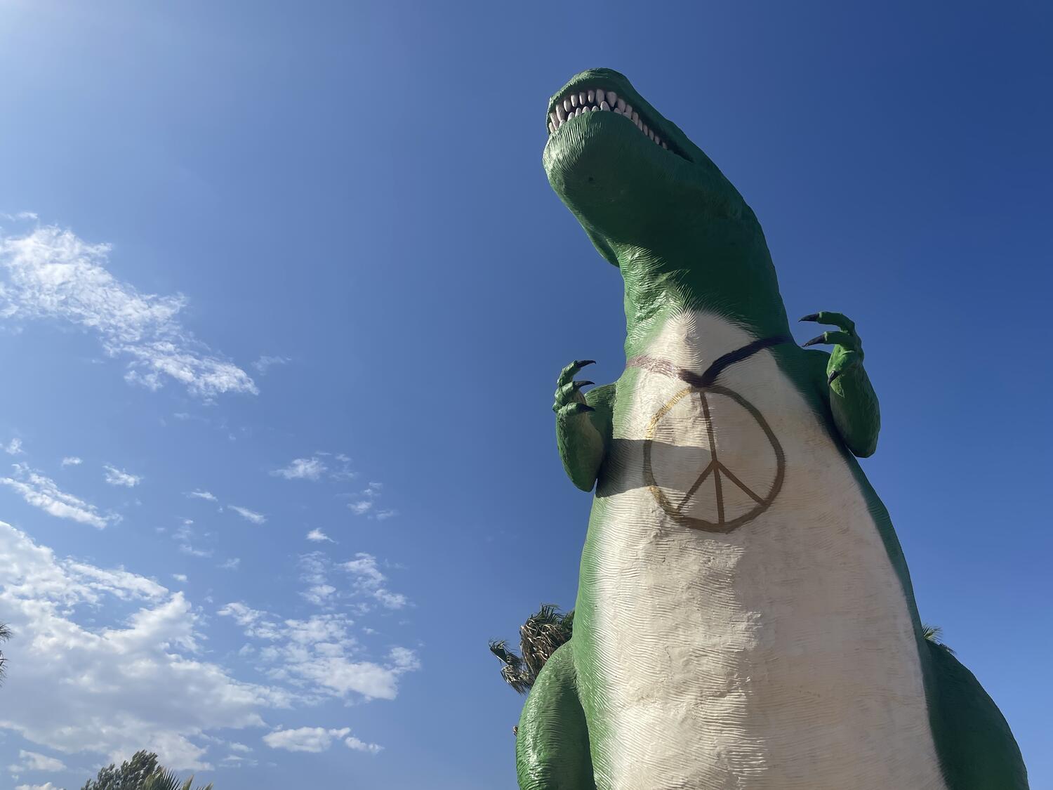 A big fiberglass t-rex seen from directly below against a blue sky. It's painted green with a white tummy, and a big peace sign necklace is painted onto it too.