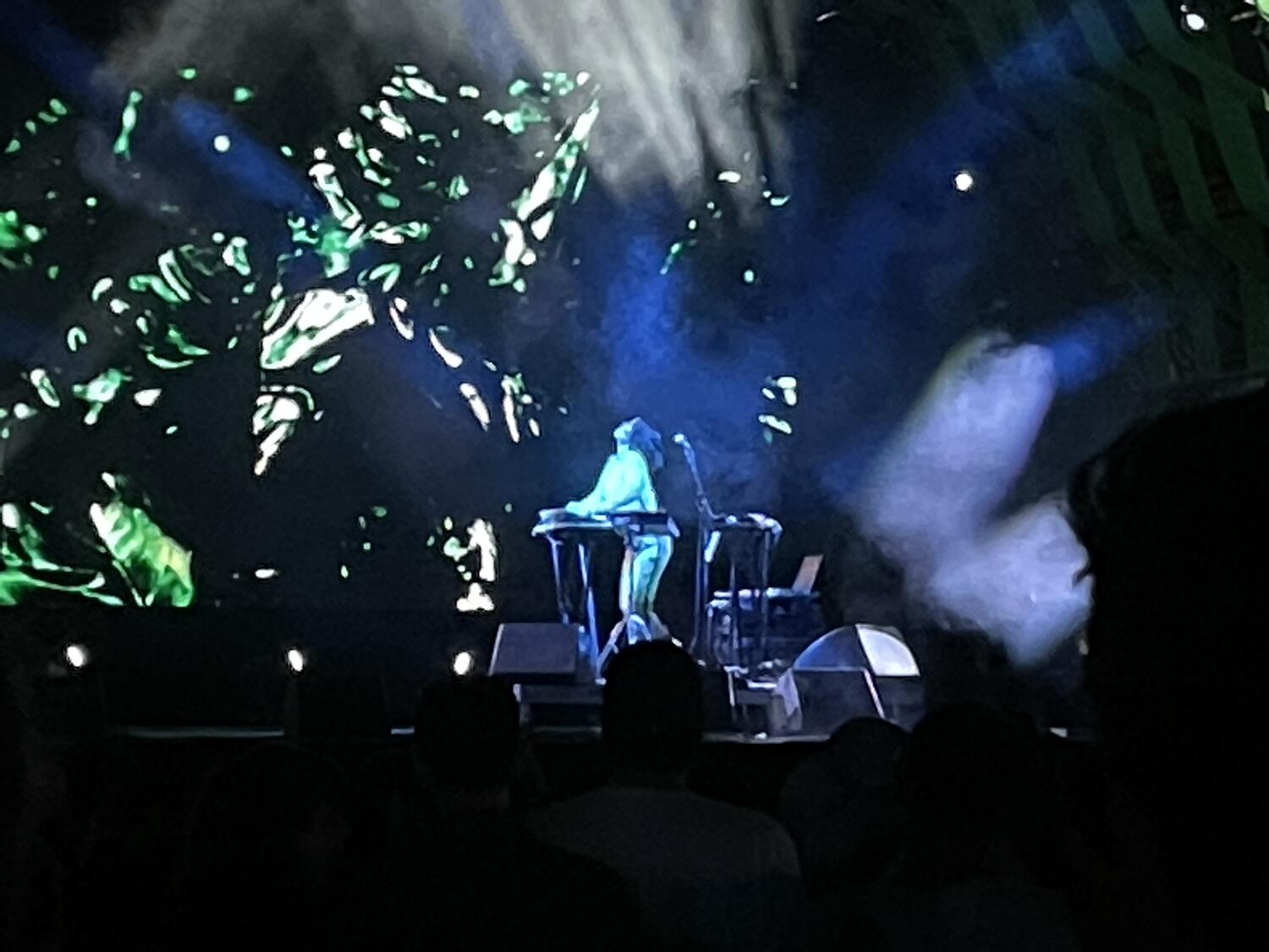 Kelly Lee Owens performing on stage. She's got two hands on a keyboard and her head is whipped back. Lots of beams of light and a colorful projection.