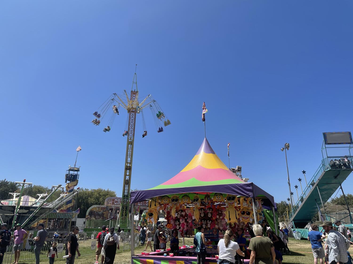 Amusement park flat rides set up in a park against a cloudless blue sky. There's also a colorful tent with some kind of game beneath it, lots of stuffed prizes hanging from the roof.