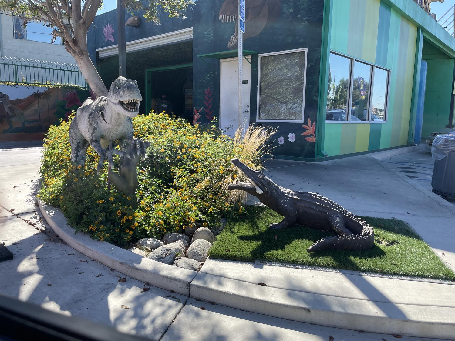 Statuses of a raptor and an alligator (?) roaring at each other in front of the entrance to a car wash. The building is painted green with more dinosaurs and prehistoric foliage decorations.