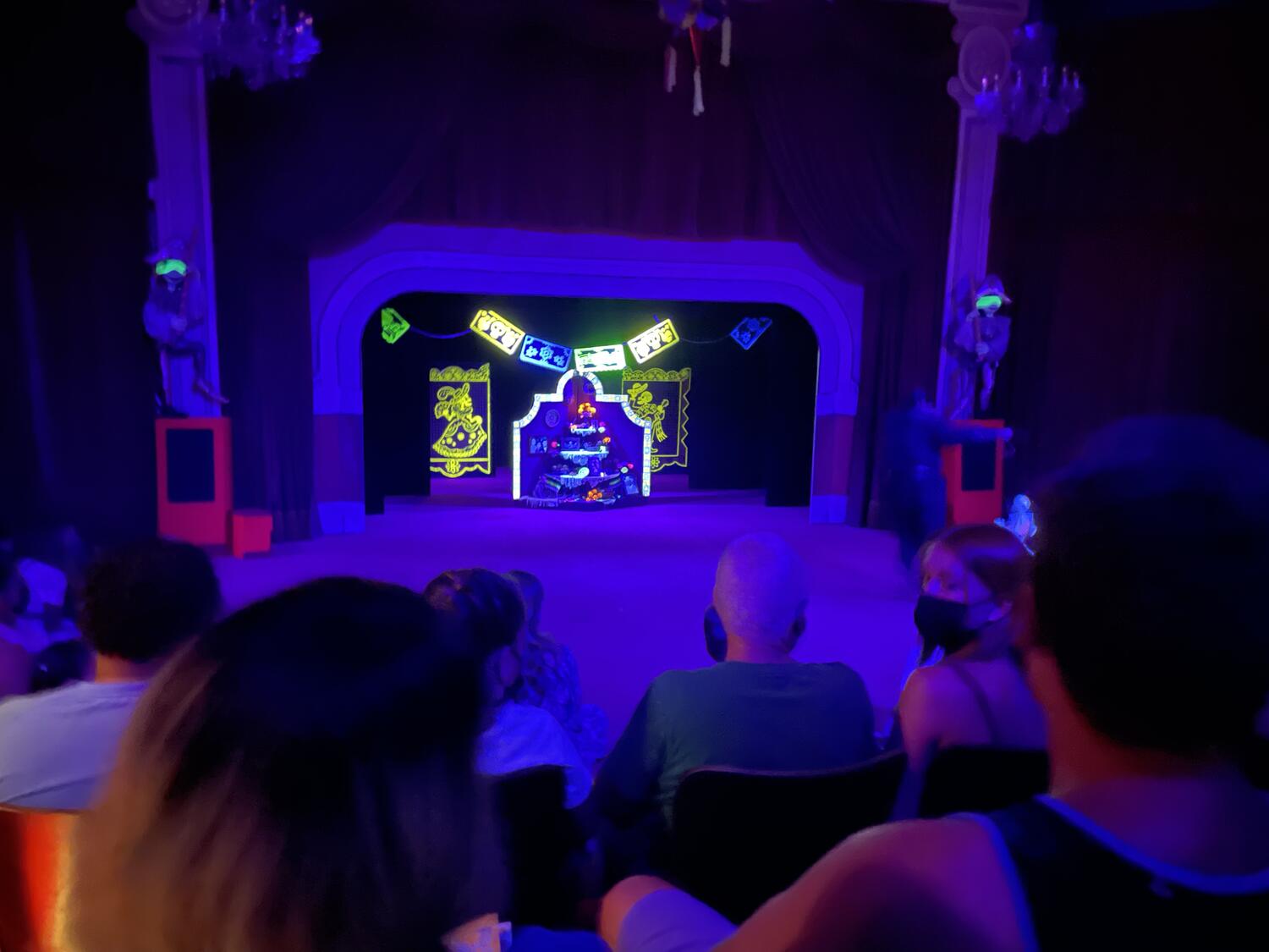 A distant shot of a puppet theater under blacklight. Glowing Day of the Dead decorations are on stage.