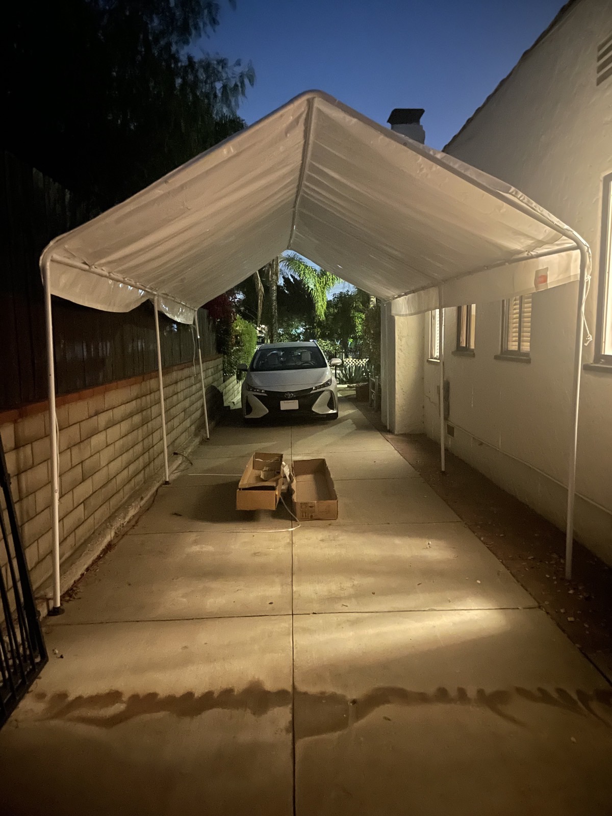 A long and narrow tent with a white tarp over its top. It's parked in the driveway with a car parked near, but not beneath it. The box it came in sits on the ground