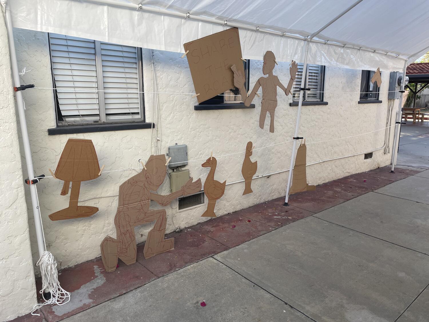 Lots of cardboard cutouts suspended under a carport tent. White clothesline runs along the poles at various heights, and the pieces are clipped to it for arrangement.