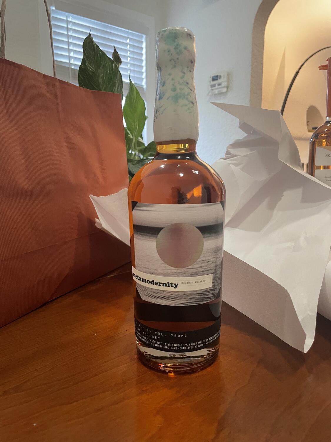 A bottle of Metamodernity whiskey. It's tall and curvy with a minimal abstract label. The need and cap are sealed with white wax that has an irregular blue and purple marbling throughout. It's surrounded by the bag and paper it was packaged with from the store.