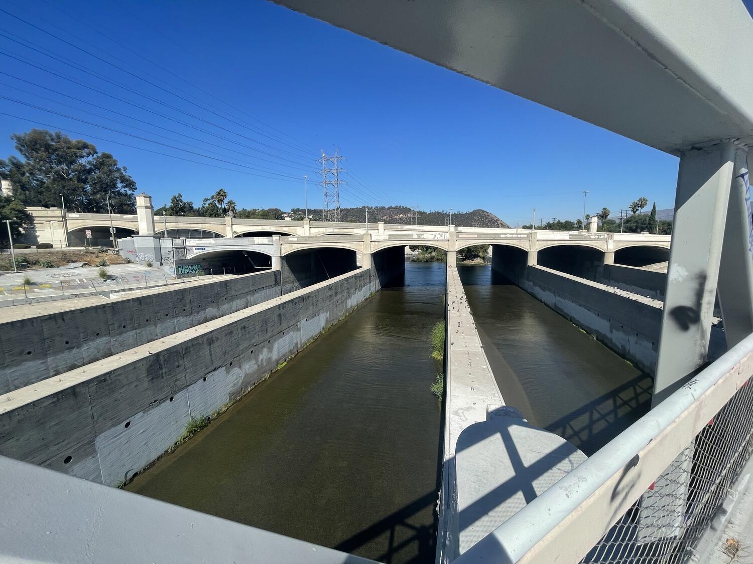 A wide-angle shot of the Glendale-Hyperion Bridge. Six arches span across the water in series, each supported by a long support wall that meets the foot bridge in the foreground (from which the picture was taken). Murky green water trickles down the avenues created by the walls.