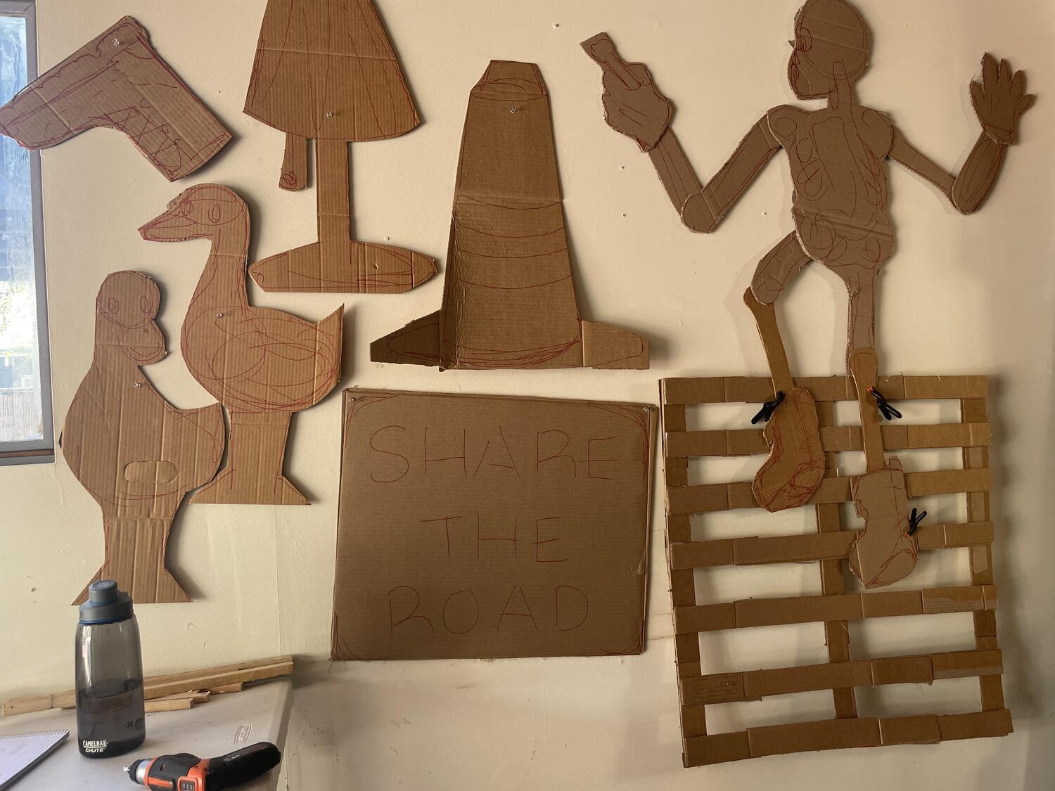 Various cardboard cutouts pinned to the wall—ducks, a boot, a lamp, a traffic cone, a street sign that reads SHARE THE ROAD, and a skeleton climbing a wooden palette with a paintbrush. There's a table against the wall with tools and supplies on it.