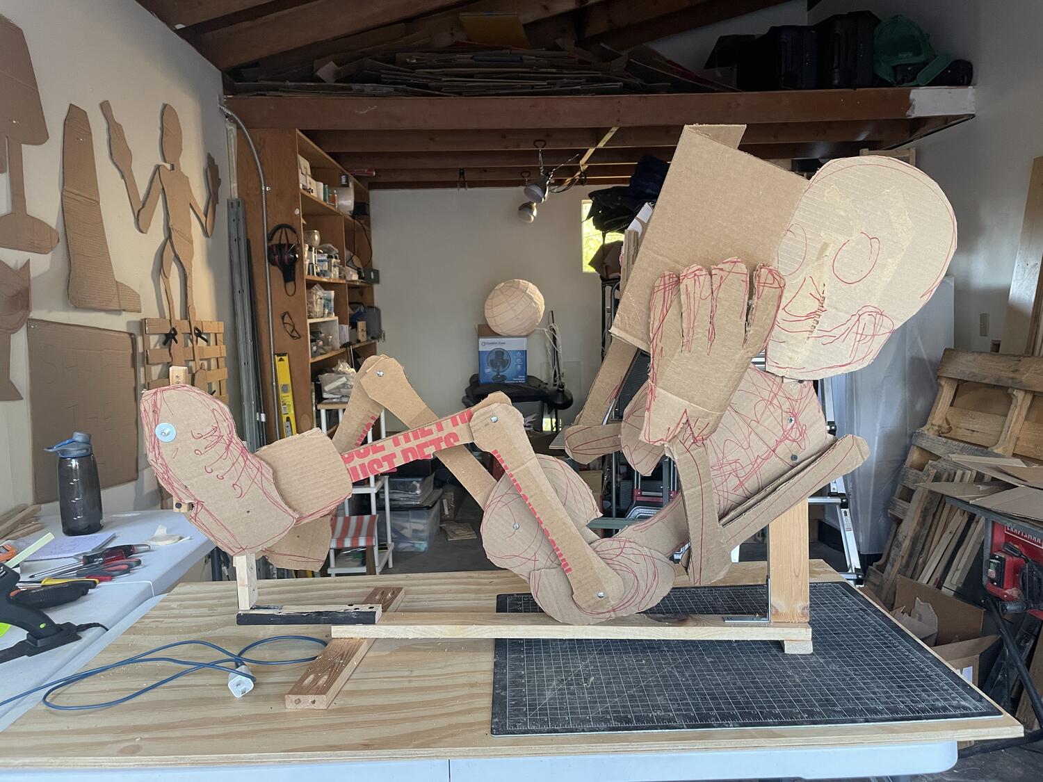 A wooden sculpture of a skeleton, reclined and holing a book up to its face. Its feet are bolted to a pedaling mechanism, like a recumbent bicycle.