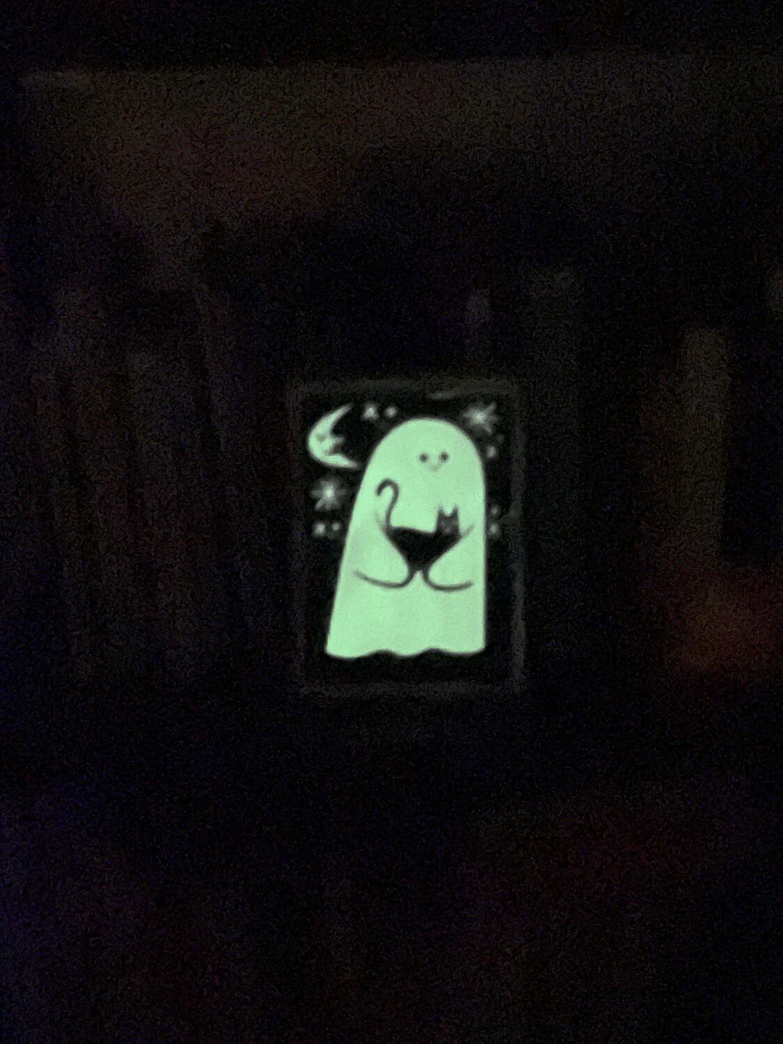 A blurry night shot of a small silkscreen print. It's a ghost holding a cat amidst the moon sand stars, and the whole thing is glowing in the dark