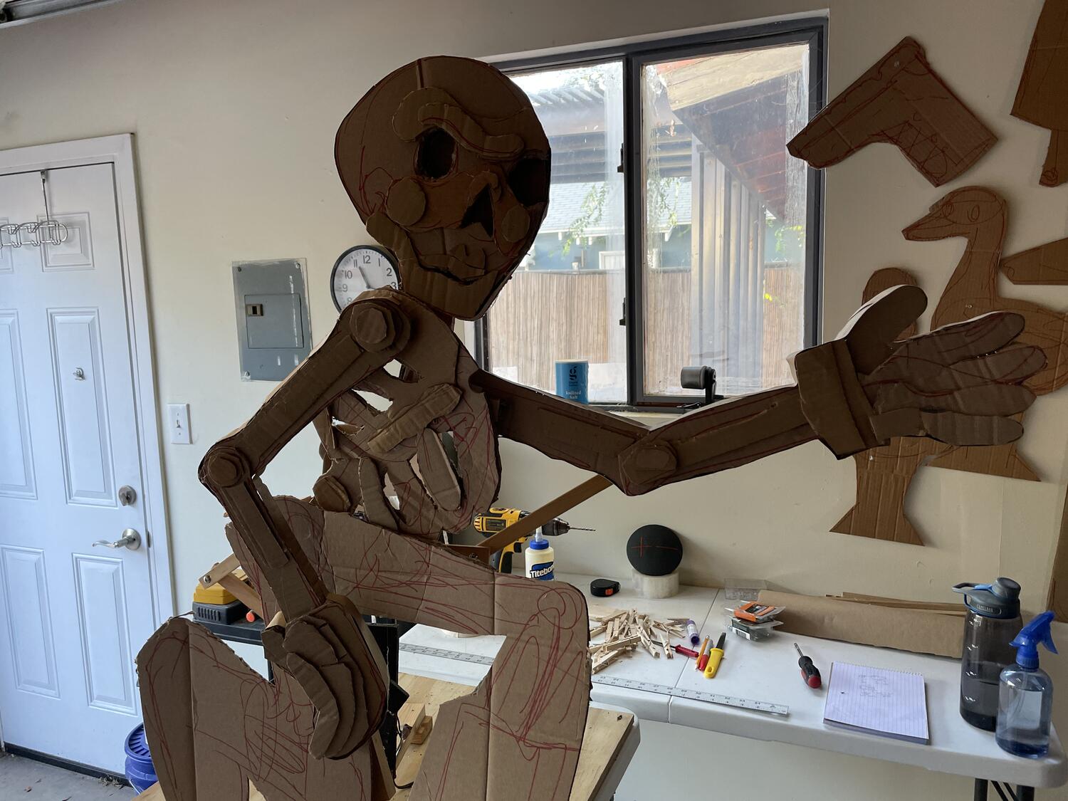 A cardboard skeleton, assembled up on a table. Tools and supplies are on another table behind it. It's kneeling down and reaching out with a gloved hand.