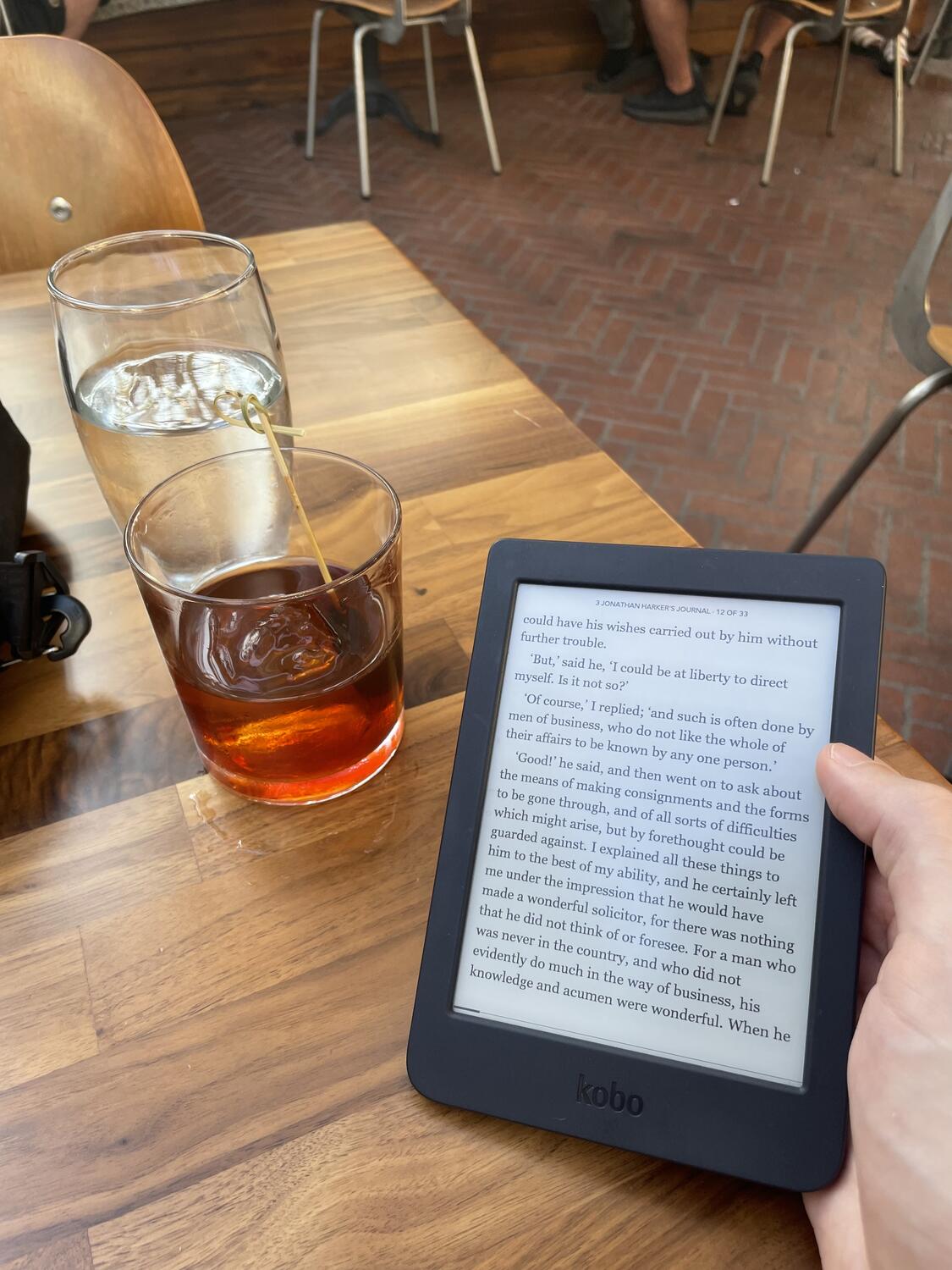 A cocktail and an e-reader resting on a nice wooden table at an outdoor restaurant. The reader is open to a page of Bram Stoker's Dracula