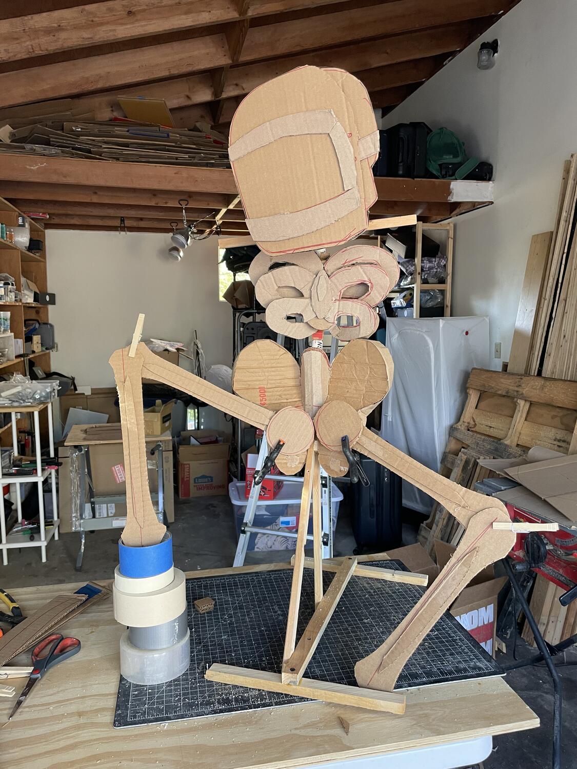 A cardboard skeleton propped up on a work table. It's got a welding mask on, and no arms yet, and its legs are just temporarily clamped in place. One leg is up on a precarious stack of rolls of tape.