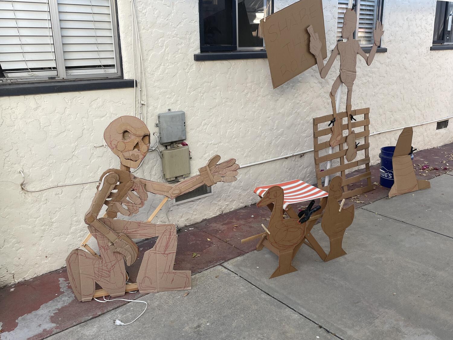 Various cardboard sculptures arranged in the driveway—the kneeling skeleton and ducks, the painter climbing up a woooden palette, a traffic cone