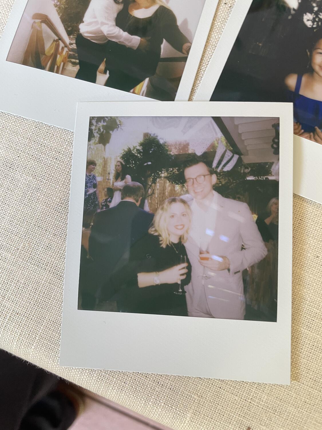 A photo of a Polaroid of me and Amy, sitting on a table among other Polaroids. We're dressed up for a wedding, smiling and holding drinks. The Polaroid is a little blurry and blown out in a really vibey way.