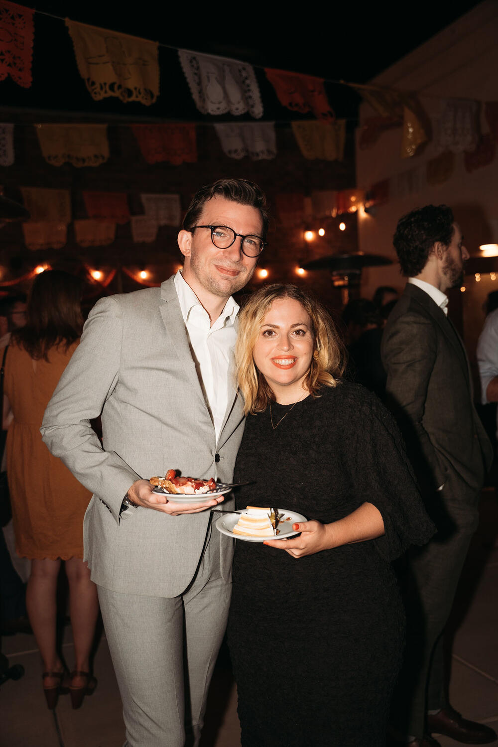 Me and Amy photographed at a wedding. I'm in a light gray suit and a crisp white shirt; she's in a dramatic black dress with fun sleeves. We're holding plates of pie and smiling at the camera. It's dark out, there are string lights, and one of those Mexican paper banners over our heads.