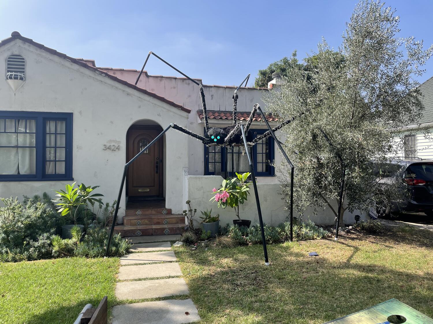 A large homemade spider decoration in front of a charming house. Its legs span from the front yard to the roof, and its body is suspended maybe seven feet off the ground. Its eyes glow green.