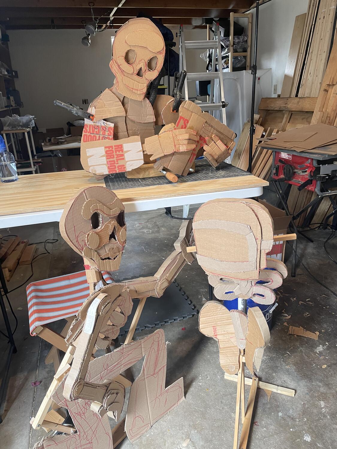 Three cardboard sculptures, nearly complete. On the ground is a kneeling skeleton beside a limbless torso in a welding mask; on the table looming above them is the bust of a skeleton in a heavy coat, holding an upturned work boot.